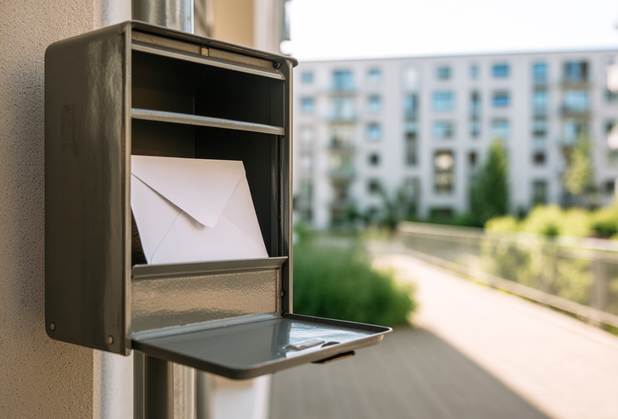 Ein weißer Briefumschlag liegt in einem Briefkasten, im Hintergrund ist ein Wohnhaus sichtbar.