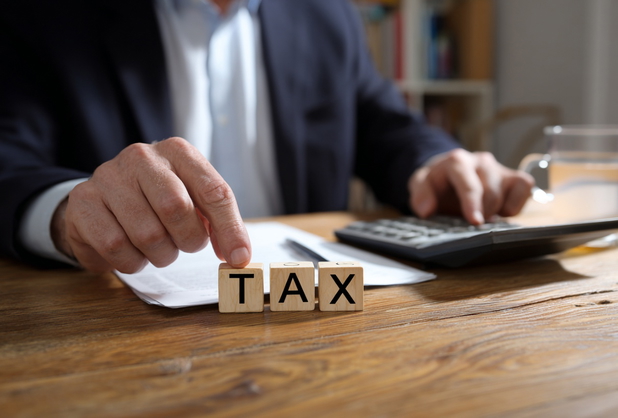 Ein Mann tippt auf einem Taschenrechner und zeigt auf drei Buchstabenwürfel "TAX".