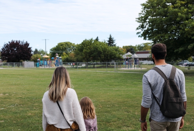 Eine junge Familie mit einer Tochter besichtigen ein Grundstück neben einem Spielplatz.