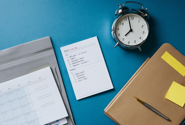 Auf einer blauen Oberfläche liegt ein Wecker, eine ToDo-Liste, ein Kalender und ein Notizbuch.
