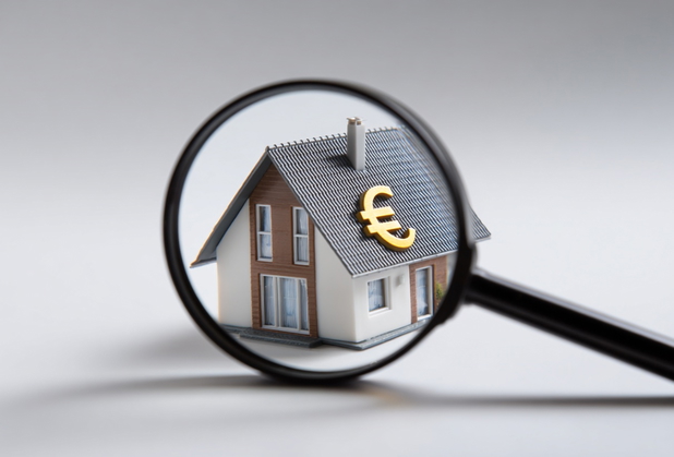 Eine Lupe untersucht ein Modellhaus, auf dessen Dach ein Euro-Zeichen abgebildet ist.