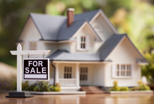 Ein Miniaturhaus auf einem Tisch mit einem "For Sale" Schild davor.