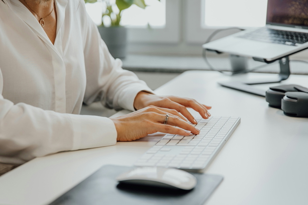 Eine Frau mit weißer Bluse sitzt am Schreibtisch und tippt auf einer weißen Mac-Tastatur.