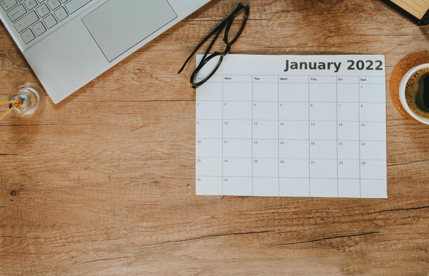 Ein Kalender, Brille, Laptop und Kaffeetasse auf einem Tisch liegend.