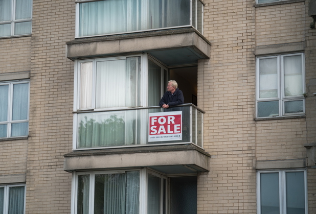 Ein Mann steht auf dem Balkon und schaut raus, an seinem Balkongelände hängt ein "For Sale"-Schild.