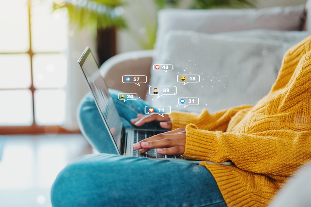 Eine Person sitzt auf einer Couch und nutzt einen Laptop, von dem mehrere Emojis ausgehen.