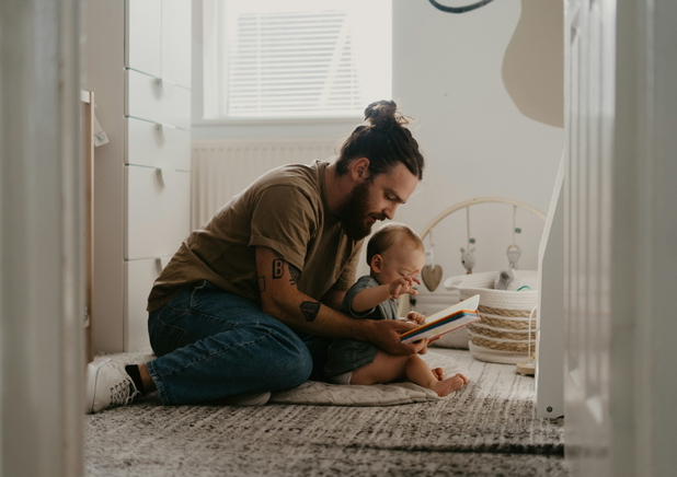 Vater sitzt mit Baby auf dem Boden und schuat ein Buch an.