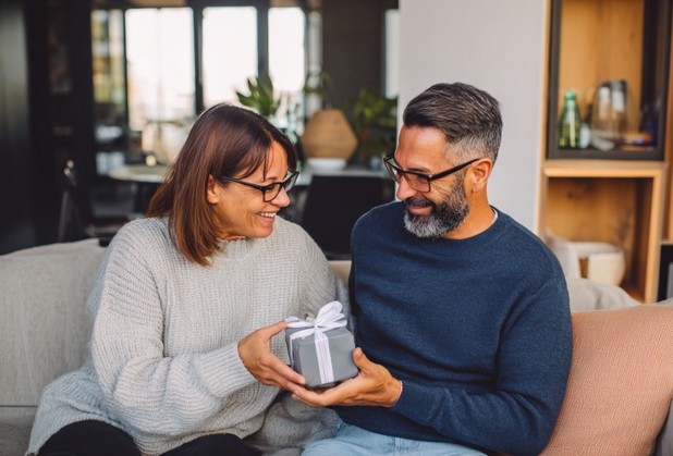 Ein Geschwisterpaar sitzt auf einer Couch, die Schwester übergibt dem Bruder ein kleines graues Geschenkpaket.