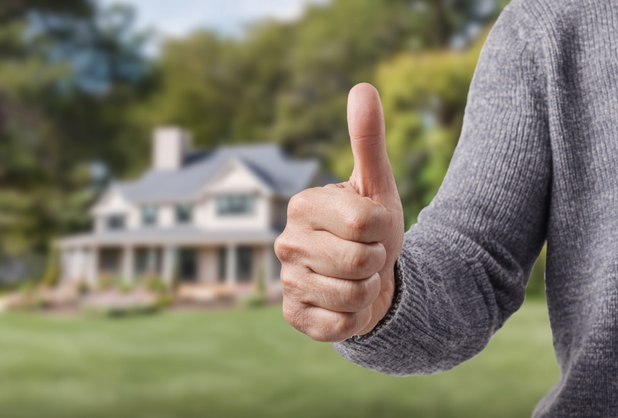 Eine Hand zeigt Daumen hoch, im Hintergrund ein Haus mit Garten.