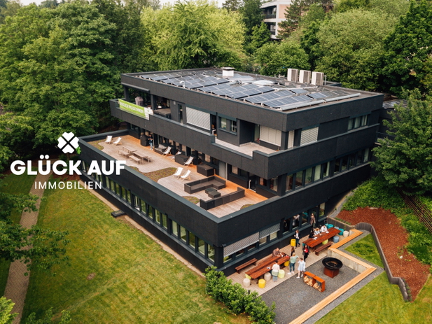 Blick auf eine moderne Villa in schwarz mit Dachterrasse und großem Garten umgeben von Glück Auf Immobilien.