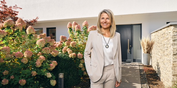Inhaberin Susanne Hagel vor einer Immobilie mir üppigem Vorgarten.