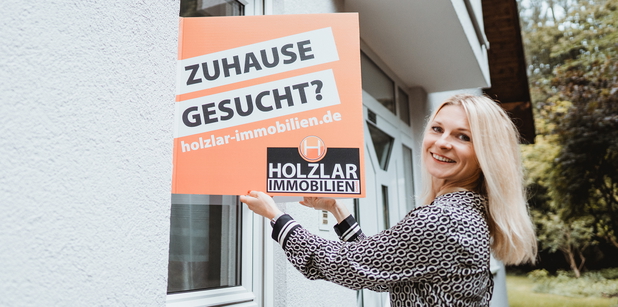 Mitarbeiterin von Holzlar Immobilien hängt ein Schild mit "Zuhause gesucht?" an einer Immobilie auf.