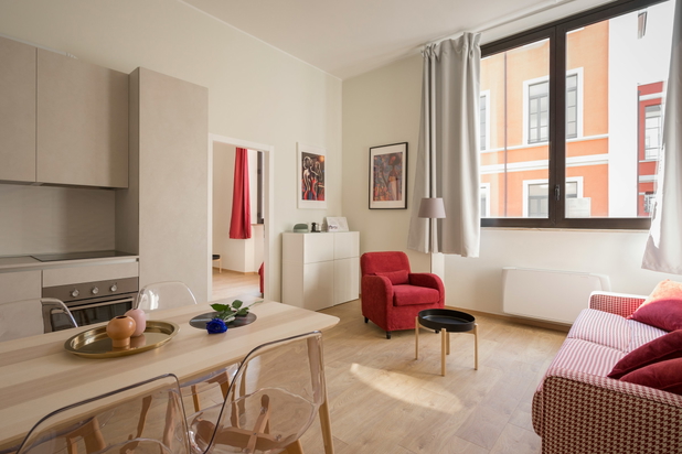 Blick in eine Wohnküche mit moderner, minimalistischer Einrichtung. Auf der rechten Bildhälfte befindet sich ein großes Fenster, vor dem ein roter Sessel steht.