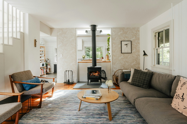 Ein Wohnzimmer mit Sofa und zwei Sesseln sowie einem Kamin, im Hintegrund ist eine Küche zu sehen.